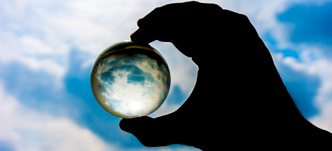 A hand holding a glass globe with a view of the clouds through it 
